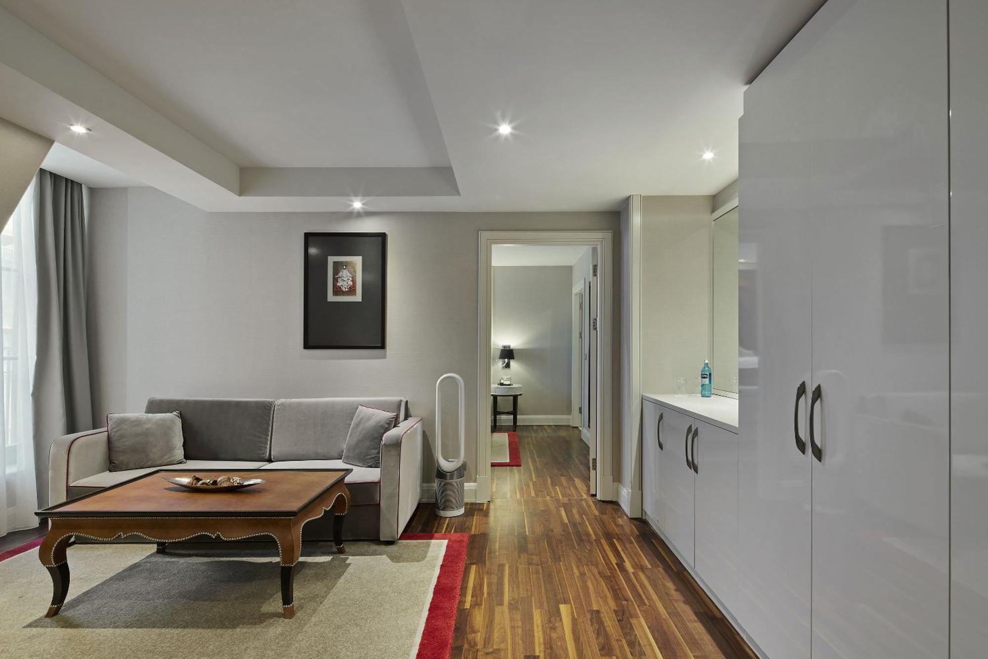 柏林御林广场泰坦尼克酒店 外观 照片 The photo shows a modern living room area featuring a gray sofa and a wooden coffee table. The floor is made of polished wood, and there is a light-colored rug underneath the table. A framed picture hangs on the wall to the left, and there are curtai