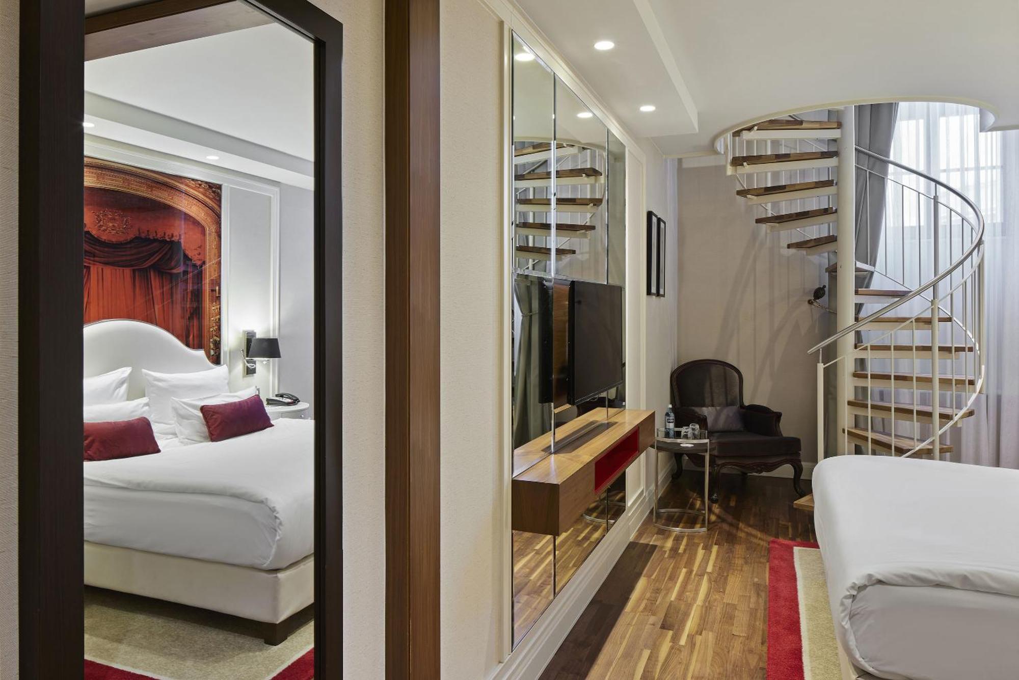 柏林御林广场泰坦尼克酒店 外观 照片 The photo shows an interior space, likely a hotel room or a suite. On one side, there is a bedroom with a large bed, decorative pillows, and artwork above the bed. The opposite side features a seating area with a dark chair and a small table, along w